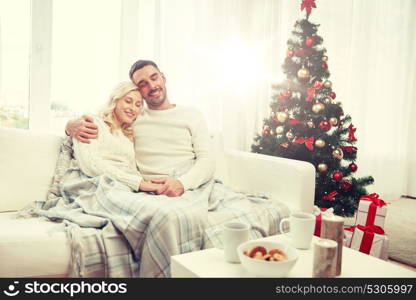 family, christmas, holidays, love and people concept - happy couple covered with plaid sitting on sofa at home. happy couple at home with christmas tree