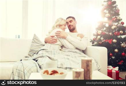 family, christmas, holidays, love and people concept - happy couple covered with plaid sitting on sofa at home. happy couple at home with christmas tree