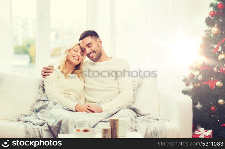 family, christmas, holidays, love and people concept - happy couple covered with plaid sitting on sofa at home. happy couple at home with christmas tree