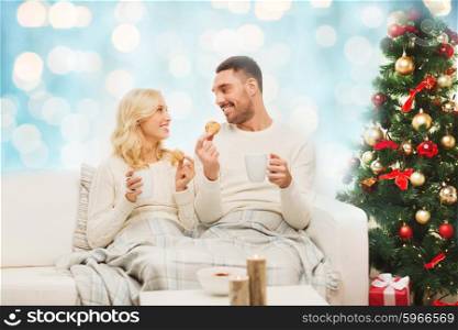 family, christmas, holidays, love and people concept - happy couple covered with plaid drinking tea and sitting on sofa at home over blue lights background