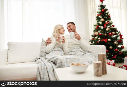 family, christmas, holidays, love and people concept - happy couple covered with plaid drinking tea and sitting on sofa at home