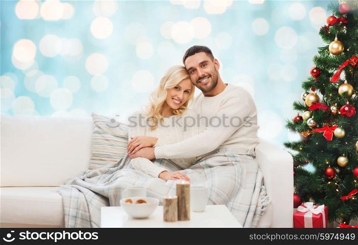 family, christmas, holidays, love and people concept - happy couple covered with plaid sitting on sofa over blue holidays lights background