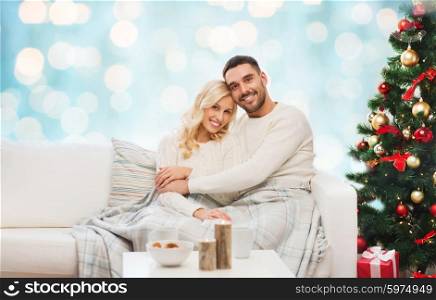 family, christmas, holidays, love and people concept - happy couple covered with plaid sitting on sofa over blue holidays lights background