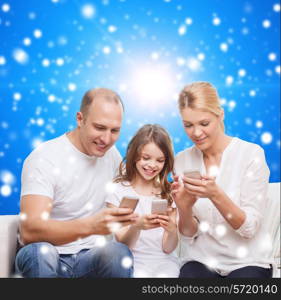 family, christmas holidays, technology and people concept - smiling mother, father and little girl with smartphones over blue snowy background