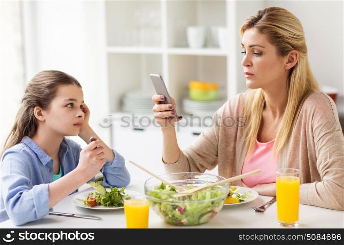 family, communication and people concept - sad girl looking at her mother with smartphone having dinner at home. sad girl looking at her mother with smartphone. sad girl looking at her mother with smartphone
