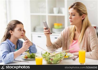 family, communication and people concept - sad girl looking at her mother with smartphone having dinner at home. sad girl looking at her mother with smartphone. sad girl looking at her mother with smartphone