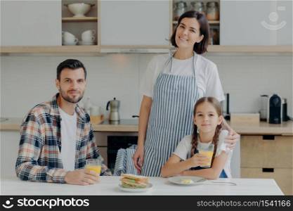Family consists of mother, father and daughter pose together in modern kitchen, drink fresh juice, eats sadwiches, enjoy domestic atmosphere. Lovely woman in apron embraces small child with love