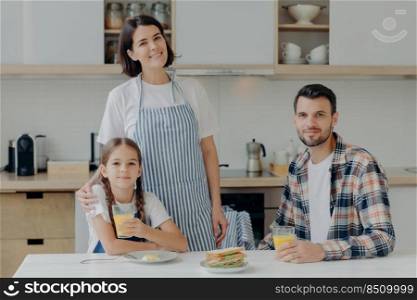 Family consists of mother, father and daughter pose together in modern kitchen, drink fresh juice, eats sadwiches, enjoy domestic atmosphere. Lovely woman in apron embraces small child with love
