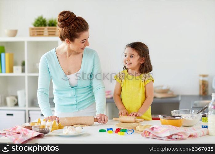 family, cooking and people concept - happy mother and little daughter with rolling pins making cookies from dough at home kitchen. happy mother and daughter making cookies at home