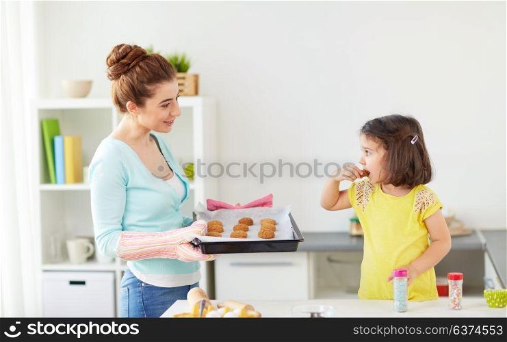 family, cooking and people concept - happy mother holding tray and little daughter eating cookie at home kitchen. happy mother and daughter baking cookies at home