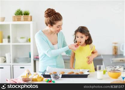 family, cooking and people concept - happy mother holding tray and little daughter eating cookie at home kitchen. happy mother and daughter baking cookies at home