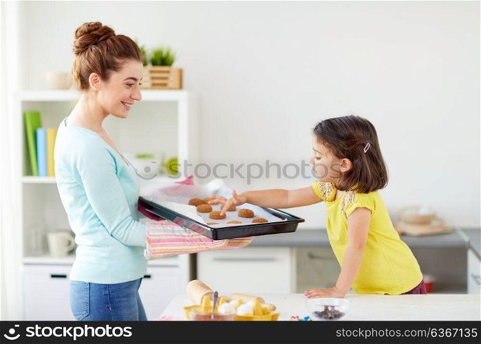 family, cooking and people concept - happy mother holding tray of cookies and little daughter at home kitchen. happy mother and daughter baking cookies at home