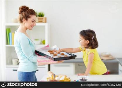 family, cooking and people concept - happy mother holding tray of cookies and little daughter at home kitchen. happy mother and daughter baking cookies at home