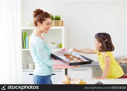 family, cooking and people concept - happy mother holding tray of cookies and little daughter at home kitchen. happy mother and daughter baking cookies at home. happy mother and daughter baking cookies at home
