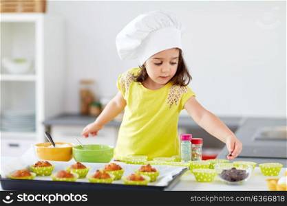 family, cooking and people concept - little girl in chefs toque baking muffins or cupcakes with sprinkles at home. little girl in chefs toque baking muffins at home