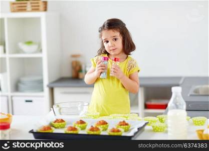 family, cooking and people concept - little girl in chefs toque baking muffins or cupcakes with sprinkles at home. little girl in chefs toque baking muffins at home