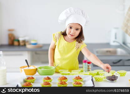 family, cooking and people concept - little girl in chefs toque baking muffins or cupcakes with chocolate sprinkles at home. little girl in chefs toque baking muffins at home