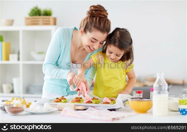 family, cooking and people concept - mother and little daughter making and decorating cupcakes with cream frosting at home kitchen. mother and daughter cooking cupcakes at home