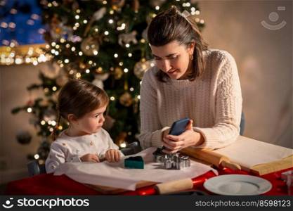 family, cooking and winter holidays concept - happy mother and baby daughter with smartphone making gingerbread cookies at home on christmas. mother and daughter making gingerbread at home