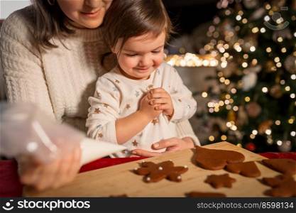 family, cooking and winter holidays concept - happy mother and baby daughter with icing in baking bag decorating gingerbread cookies at home on christmas. mother and daughter decorating gingerbread at home