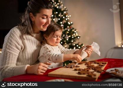 family, cooking and winter holidays concept - happy mother and baby daughter with icing in baking bag decorating gingerbread cookies at home on christmas. mother and daughter decorating gingerbread at home