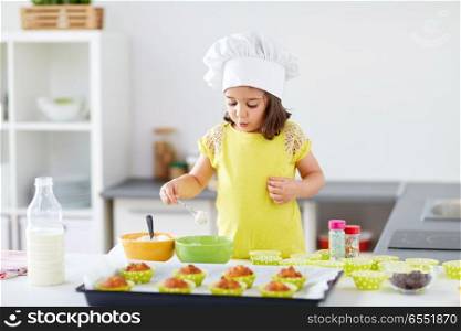family, cooking, baking and people concept - little girl in chefs toque making batter for muffins or cupcakes at home kitchen. little girl in chefs toque baking muffins at home. little girl in chefs toque baking muffins at home