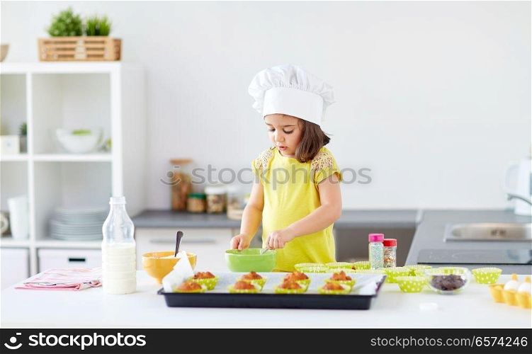 family, cooking, baking and people concept - little girl in chefs toque making batter for muffins or cupcakes at home kitchen. little girl in chefs toque baking muffins at home