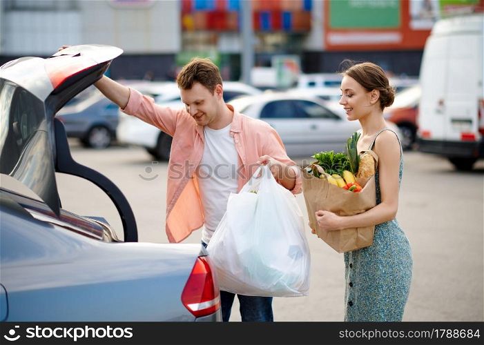 Family couple puts their purchases in the trunk on supermarket car parking. Happy customers carrying purchases from the shopping center, vehicles on background. Family couple puts their purchases in the trunk