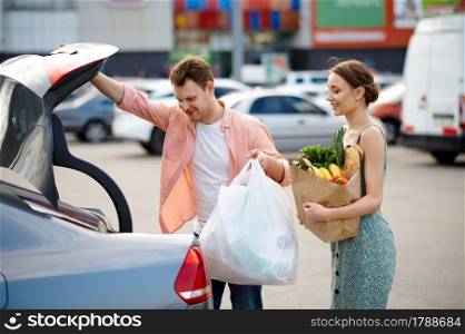 Family couple puts their purchases in the trunk on supermarket car parking. Happy customers carrying purchases from the shopping center, vehicles on background. Family couple puts their purchases in the trunk