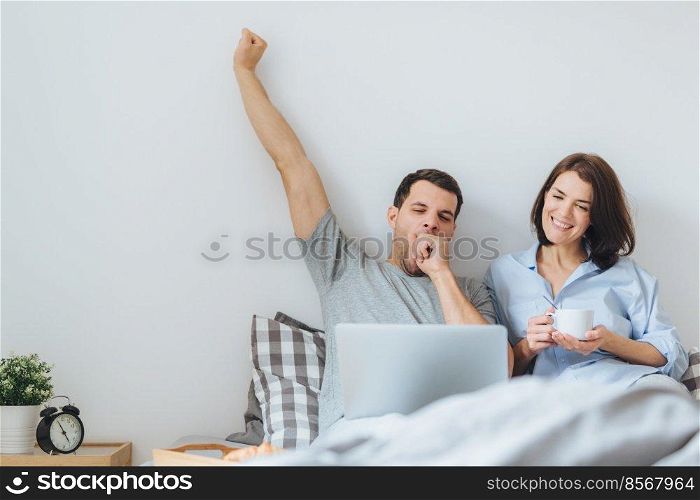 Family couple spend weekends together in bedroom, watch video or films, drink coffee. Handsome man stretches and yawns as watches boring film with his wife. People, relaxation, family concept