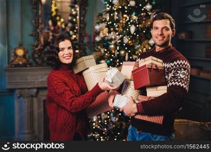 Family couple stand near decorated New Year or Christmas tree, hold many presents, being busy with holiday preparations, have positive expressions. Young woman and man prepare for celebration