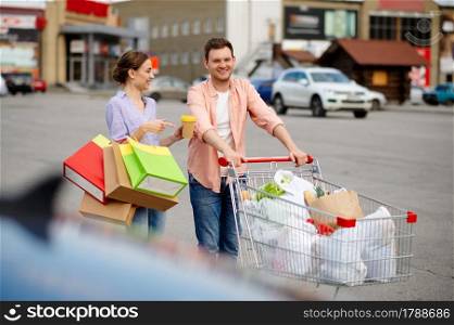 Family couple with bags in cart on supermarket car parking. Happy customers carrying purchases from the shopping center, vehicles on background. Family couple with bags in cart on car parking