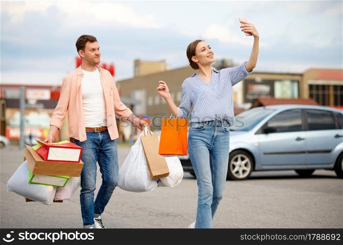 Family couple with cardboard bags makes selfie on supermarket car parking. Happy customers carrying purchases from the shopping center, vehicles on background. Family couple with bags makes selfie on parking