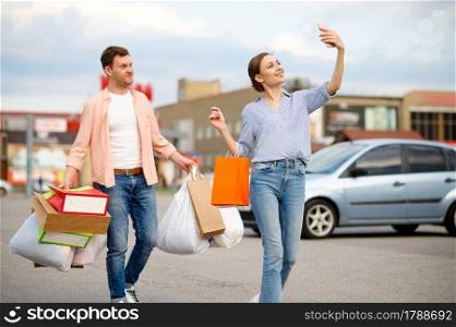 Family couple with cardboard bags makes selfie on supermarket car parking. Happy customers carrying purchases from the shopping center, vehicles on background. Family couple with bags makes selfie on parking