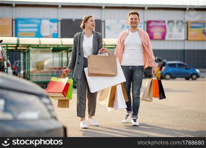 Family couple with cardboard bags on supermarket car parking. Happy customers carrying purchases from the shopping center, vehicles on background. Family couple with bags on supermarket parking