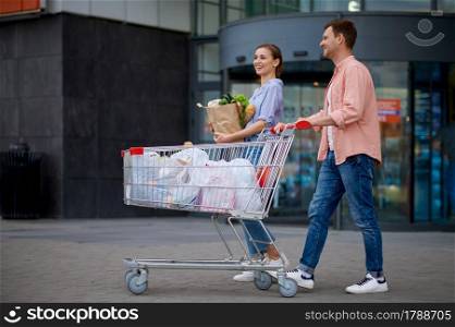 Family couple with cart on market car parking. Cheerful customers carrying purchases from the shopping center. Family couple with cart on market car parking