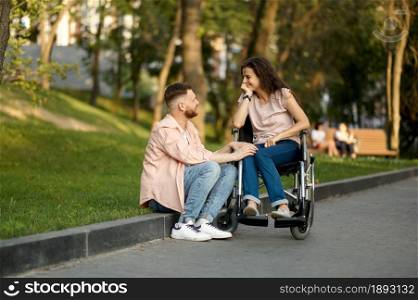 Family couple with wheelchair leisures in park. Paralyzed people and disability, care for a disabled woman. Husband and handicapped wife overcome difficulties together, warm relationships. Couple with wheelchair in park, disabled woman
