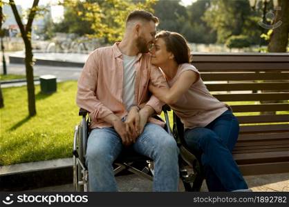 Family couple with wheelchair sitting on bench in park. Paralyzed people and disability, care for a disabled man. Husband and wife overcome difficulties together, warm relationships. Couple with wheelchair sitting on bench in park