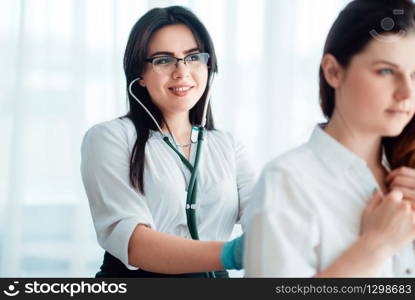 Family doctor listens to female patient using a stethoscope, reception in clinic. Specialist examines woman, professional health care. Doctor listens to patient using a stethoscope