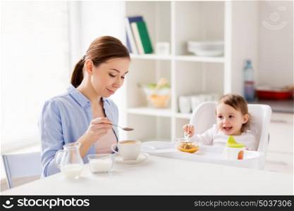 family, eating, breakfast and people concept - happy mother adding sugar to coffee cup and little baby with food sitting in highchair at home kitchen. happy mother and baby having breakfast at home