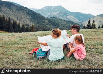 Family examining a map of mountains trials sitting on grass enjoying summer day during vacation trip in mountains. People actively spending time outside. Mountain landscape in the background. Family examining a map of mountains trials sitting on grass enjoying summer day during vacation trip in mountains