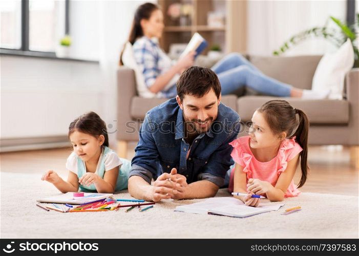 family, fatherhood and leisure concept - father spending time with his little daughters drawing in sketchbooks by crayons and lying on floor at home. father with little daughters drawing at home