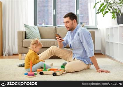 family, fatherhood and people concept - father with smartphone and little baby daughter playing with wooden toy toy blocks kit at home. father with smartphone and baby daughter at home
