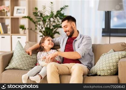 family, fatherhood and people concept - happy father and daughter eating popcorn on sofa at home. happy father and daughter eating popcorn at home