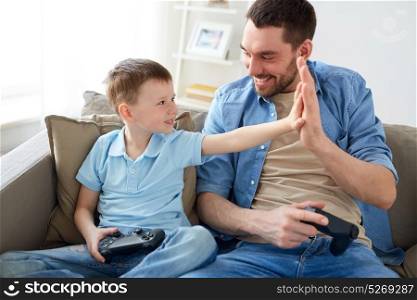 family, fatherhood and people concept - happy father and little son with gamepads playing video game and doing high five at home. father and son with gamepads doing high five