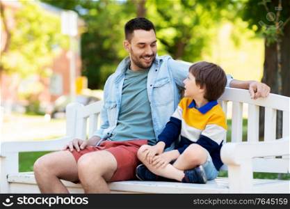 family, fatherhood and people concept - happy father and son sitting on bench at summer park and talking. father with son sitting on park bench and talking