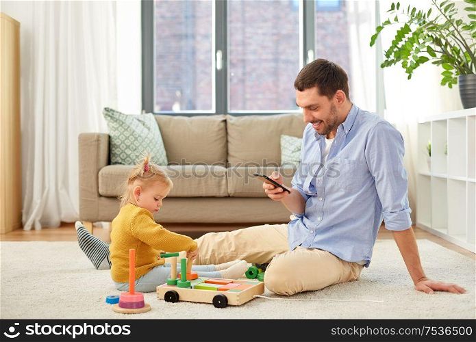 family, fatherhood and people concept - happy father with smartphone and little baby daughter playing with wooden toy toy blocks kit at home. father with smartphone and baby daughter at home