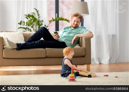 family, fatherhood and people concept - happy red haired father with smartphone and little baby daughter playing with toy blocks kit at home. father and baby playing with toy blocks at home