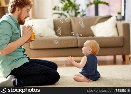 family, fatherhood and people concept - red haired father and little baby daughter playing with soap bubbles at home. father and baby daughter with soap bubbles at home