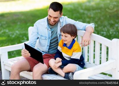 family, fatherhood and technology concept - happy father and little son with tablet pc computer sitting on bench at summer park. father and son with tablet pc computer at park